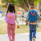 Two children walking in park wearing lunch totes on their backs. 